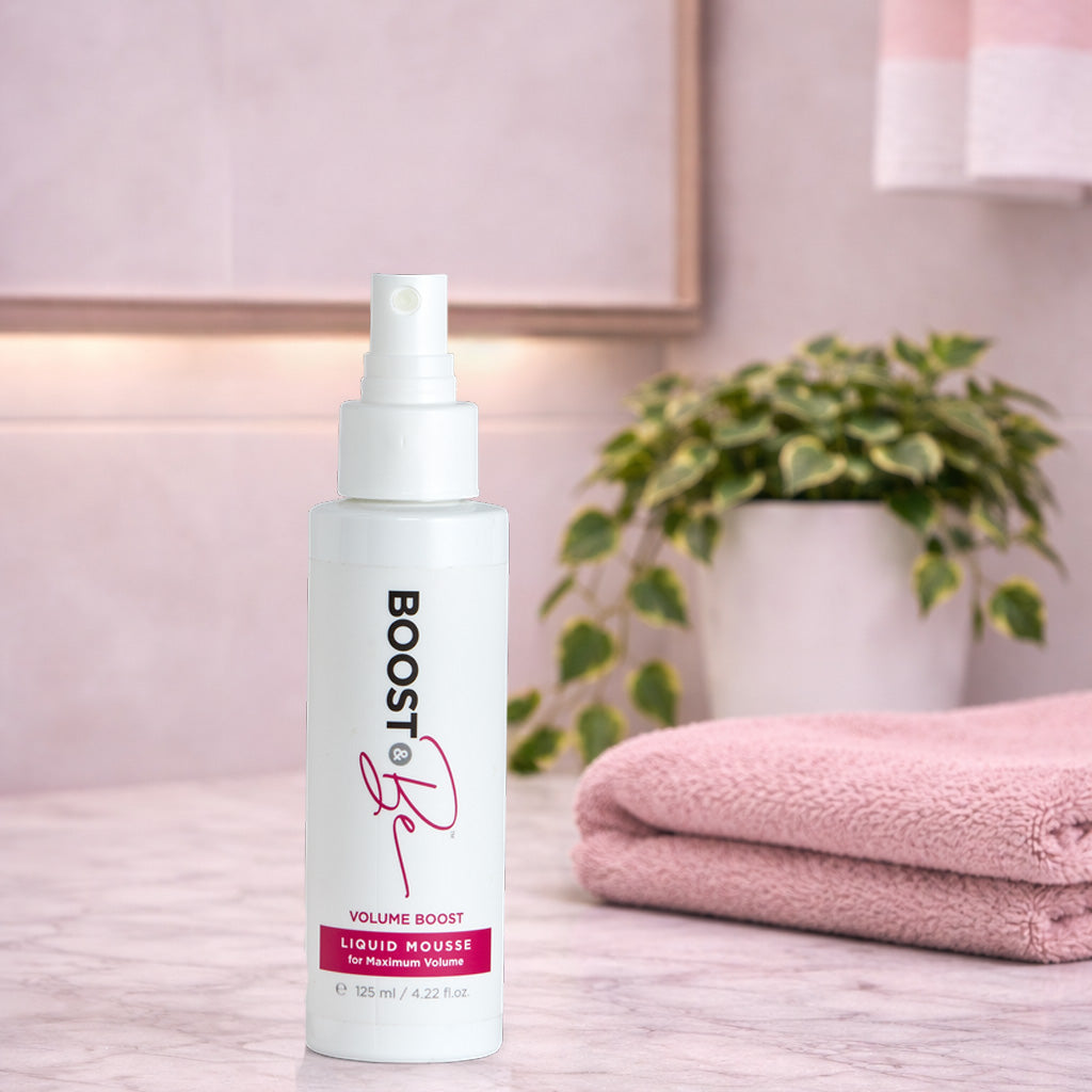Boost & Be Volume Boost Liquid Mousse bottle on a bathroom counter with towels and a plant in the background.